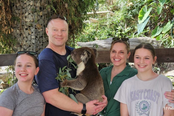 Four people smiling with a koala in a lush outdoor setting.
