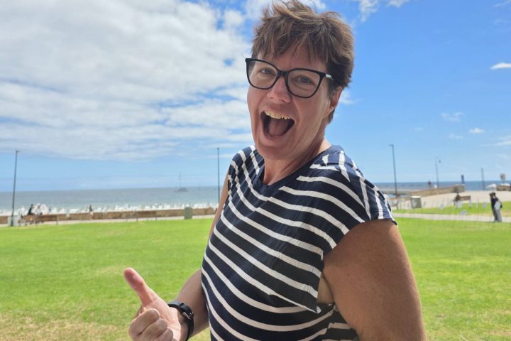 Smiling person in striped shirt giving thumbs up near a beach under a cloudy blue sky.