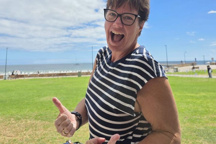 Smiling person in striped shirt giving thumbs up near a beach under a cloudy blue sky.