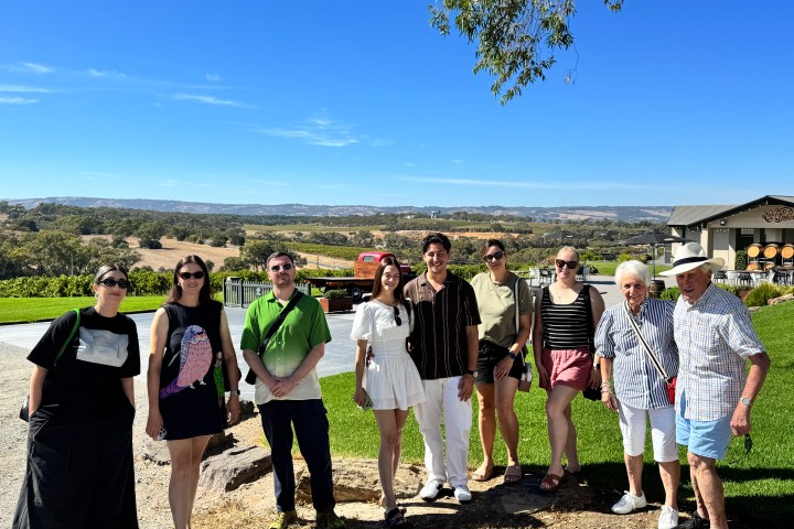 Group of people posing outside with vineyard and blue sky in background.