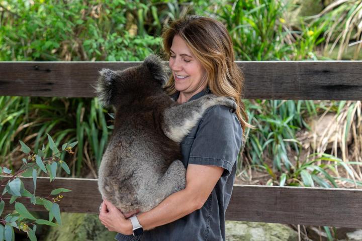 Woman holding a koala bear with a smile, standing in front of greenery.