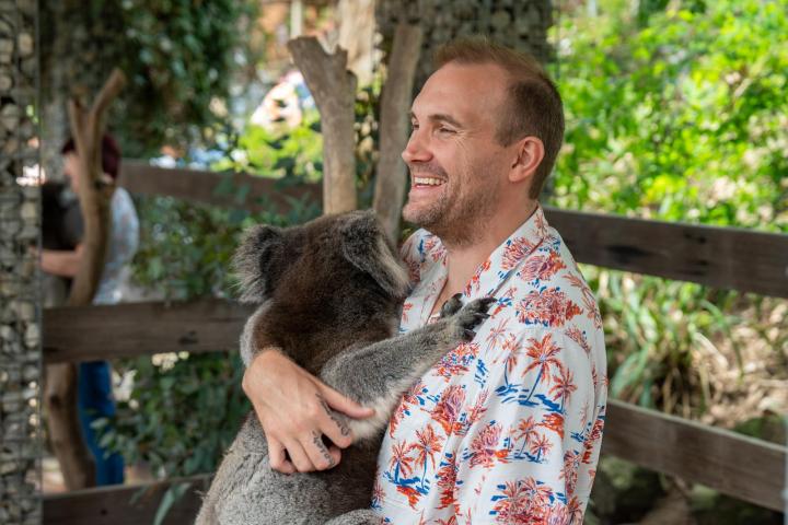 Smiling man in a floral shirt holding a koala in an outdoor setting.
