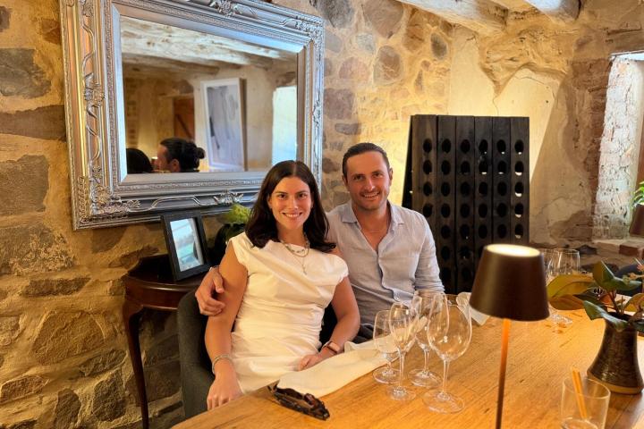 Couple seated at rustic table with empty wine glasses in cozy stone-walled room with mirror.