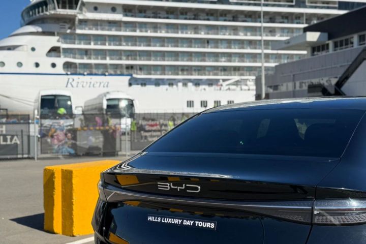 Black BYD car parked near a large cruise ship in a sunny port.