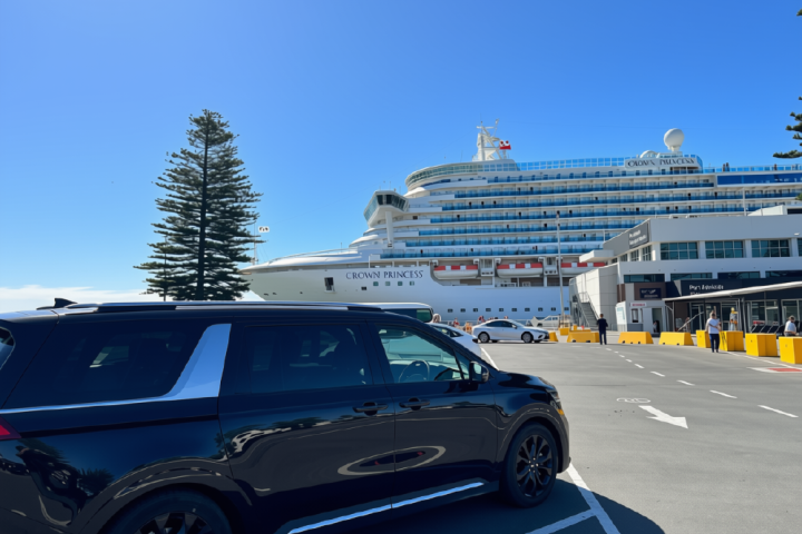 Black SUV parked near a large cruise ship and terminal under a clear blue sky.