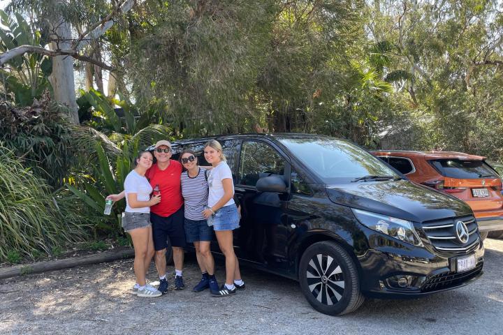 Four people smiling by a black Mercedes van in a sunny parking area.