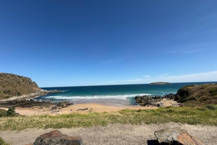 Coastal landscape with sandy beach, rocky cliffs, and an island under clear blue sky.