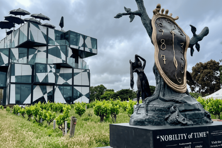 Surreal clock sculpture titled 'Nobility of Time' near modern, geometric building in vineyard setting.