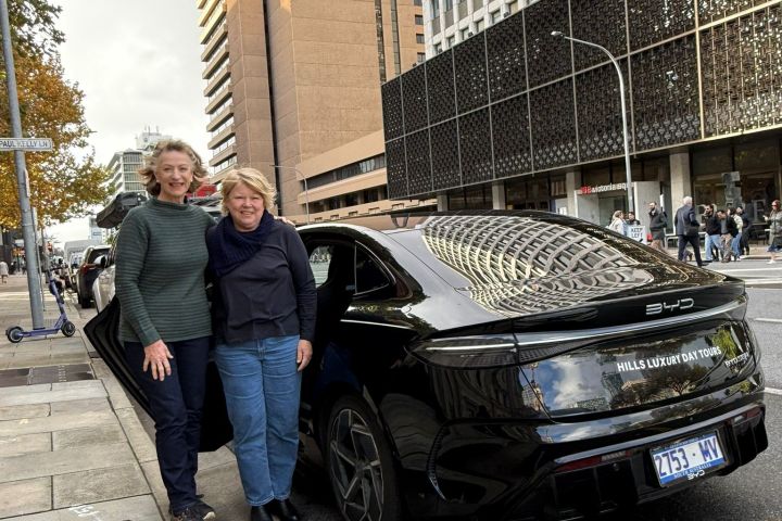 Two people stand by a parked black car on a city street with tall buildings.